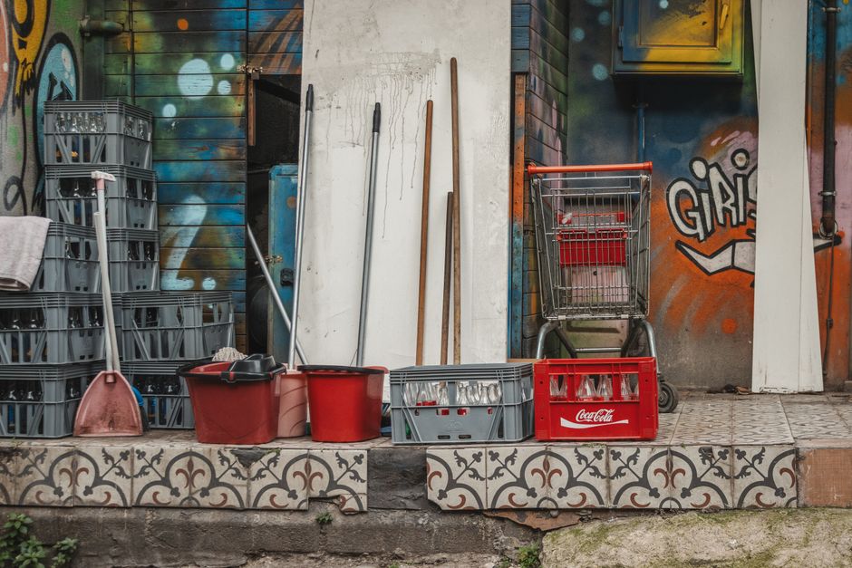 An outdoor urban scene featuring cleaning tools and supplies arranged on the pavement in front of a graffiti-covered wall. The items include stacked gray plastic crates, a pink scoop, two red buckets,