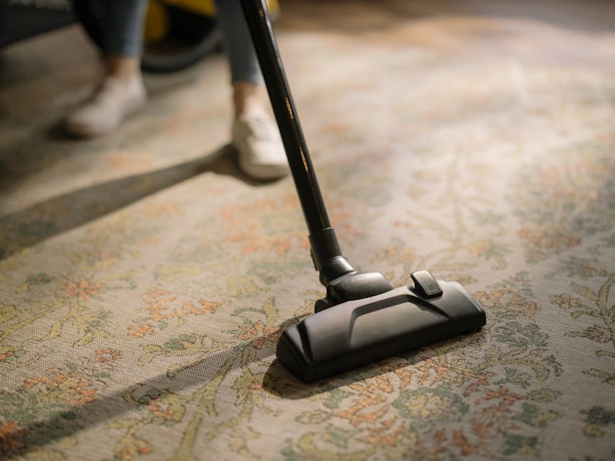 A woman using a yellow vacuum cleaner to deep clean a patterned area rug in a living room, featuring a hardwood floor underneath. The scene is well-lit, with natural lighting illuminating the space, a