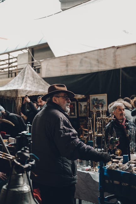 A man with a dark jacket, hat, and glasses is standing behind a stall at Brixton Market, surrounded by various household items and decorative objects. The stall features a table draped with a white cl