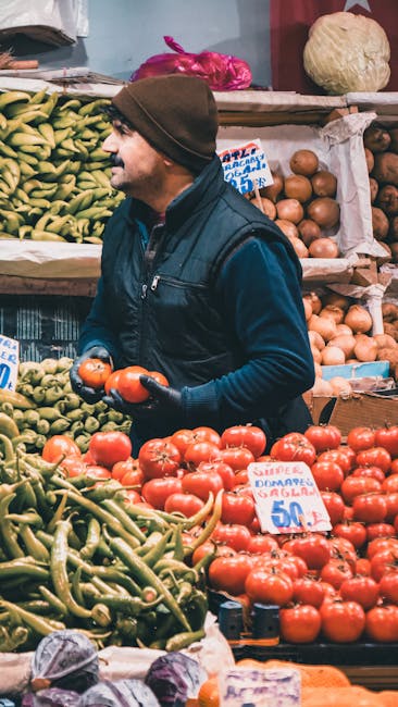A man with a dark jacket, hat, and glasses is standing behind a stall at Brixton Market, surrounded by various household items and decorative objects. The stall features a table draped with a white cl